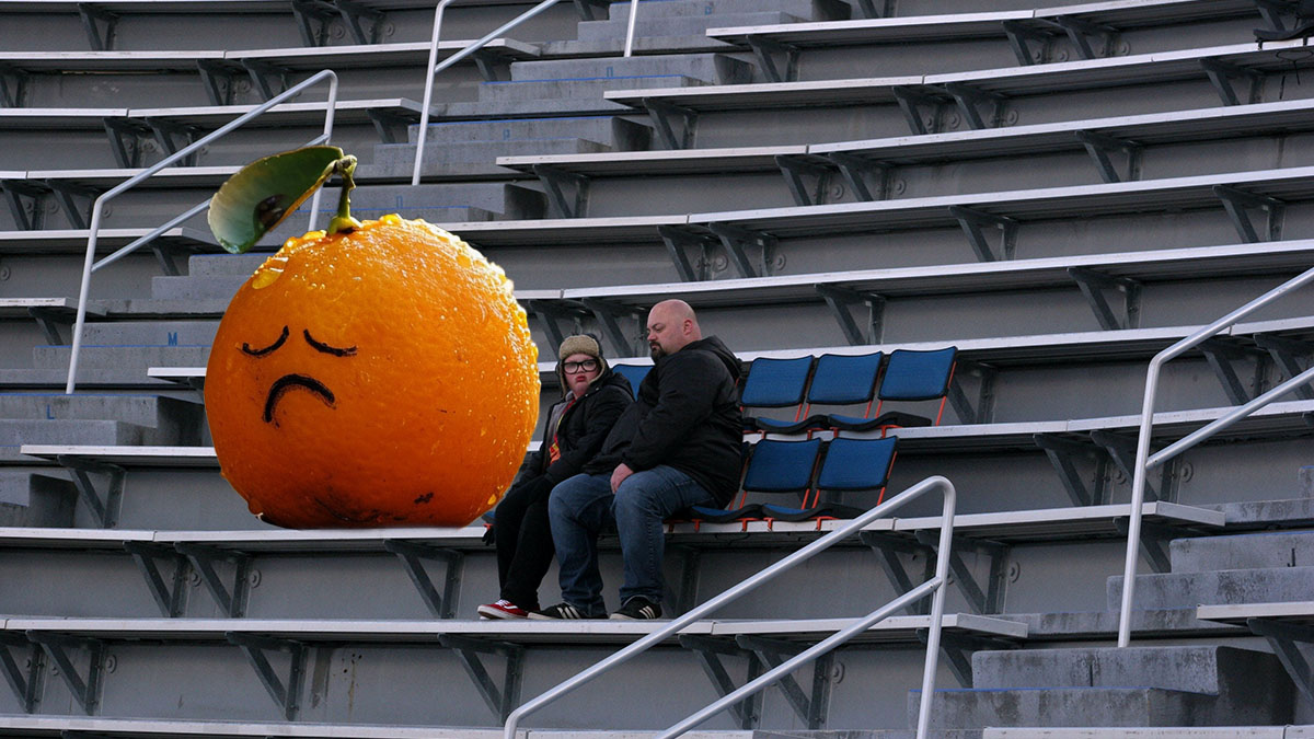 Two people sitting in bleachers in alone in an empty football stadium next to a crying orange.